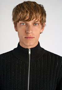 Schwarzer Strickpullover mit hohem Kragen und Reißverschluss, der ein strukturiertes Muster und gerippte Akzente aufweist, vor einem neutralen Hintergrund präsentiert.