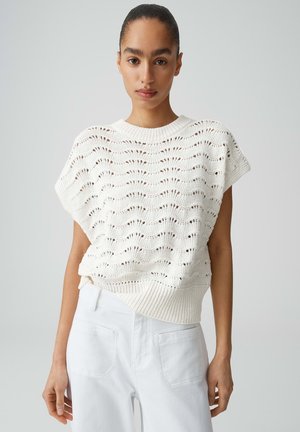 Vrouw draagt een mouwloos wit gebreid topje met golfpatronen en witte broek, staand tegen een effen lichte achtergrond.