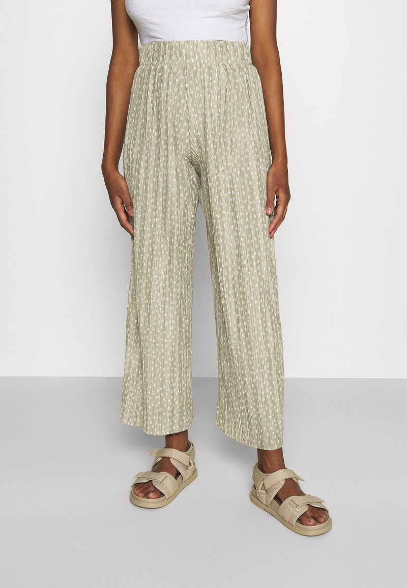 Personne portant un pantalon large plissé vert clair à petit motif blanc, un haut blanc et des sandales beiges, debout à l'intérieur.