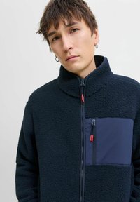 Mornarska fleece jakna s visokim ovratnikom, zatvorenim prednjim dijelom i džepom na prsima. Tekstura tkanine s glatkim panelima u akcentskim bojama i crvenim patentnim zatvaračima.