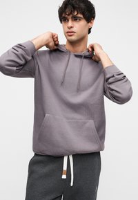 Γκρι φούτερ με κουκούλα στο μπροστινό μέρος, κορδόνι στην κουκούλα και ριμπ μανσέτες, συνδυασμένο με γκρι φόρμα με μικρή ετικέτα.