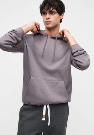 Hanorac gri cu buzunar frontal, glugă cu șnur și manșete striate, asortat cu pantaloni de trening gri cu o etichetă mică.