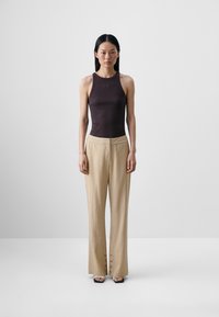 Brun uten ermer bodysuit kombinert med beige, teksturerte bukser med vide ben som har knapper på kanten. Brukt med svarte flate sandaler.