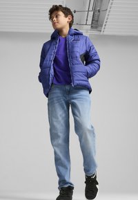 Blå pufferjakke med hætte, der har kviltet tekstur og grå accenter, båret over en lilla skjorte, parret med lyseblå jeans og sorte sneakers.
