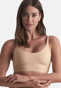 Donna con lunghi capelli castani che indossa un reggiseno color carne senza cuciture, con le braccia incrociate davanti al torso su uno sfondo neutro.