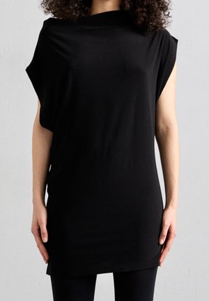 Woman with curly hair wearing a sleeveless black tunic top and black leggings against a plain gray background.