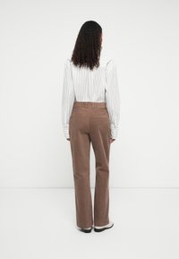 Persoon met krullend haar, gekleed in een wit gestreept shirt, bruine corduroybroek en lichtgekleurde schoenen, staand met het gezicht naar een witte achtergrond.