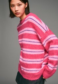 Růžovo-fialový pruhovaný svetr vyrobený z jemného, chlupatého materiálu, s kulatým výstřihem a volným střihem, nošený s tmavými džíny.