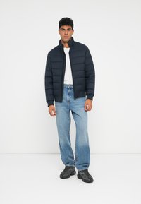 Jachetă puffer bleumarin cu guler înalt, închidere cu fermoar și design matlasat, purtată cu un tricou alb și blugi albaștri deschiși. Pantofi sport negri.