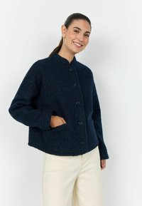 Geacă texturată bleumarin cu guler în picioare, închidere frontală cu nasturi și buzunare laterale. Material moale și croială lejeră, asortată cu pantaloni deschiși la culoare.