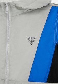 Svetlo siva jakna z modrimi in črnimi akcenti, ki ima zadrgo in trikotno logotipno značko na prsih. Gladka tekstura.