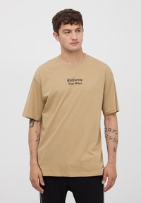 Beżowa bawełniana koszulka z krótkimi rękawami, z napisem "California Never Stops" wydrukowanym na czole czarnym kolorem. Swobodny, luźny krój.