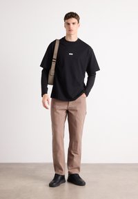 T-shirt nera a maniche corte con logo, indossata sopra una maglietta nera a maniche lunghe. Pantaloni marrone chiaro con tasche e scarpe nere con lacci.