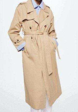 Trench-coat beige à double boutonnage, taille ceinturée et poignets boutonnés porté sur une chemise rayée bleue avec les mains dans les poches.