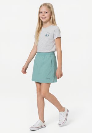 Jente med langt blond hår som har på seg en grå t-skjorte, et blågrønt skjørt og hvite joggesko, går med et lett smil på en hvit bakgrunn.