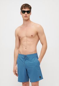 Blaue Badeshorts aus leichtem, glattem Stoff mit elastischem Bund und einer seitlichen Tasche. Das Model trägt schwarze Sonnenbrillen.