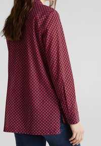 Camicia rossa con maniche lunghe, caratterizzata da un motivo circolare ripetuto in blu e bianco. Lunghezza tunica con spacchi laterali, realizzata in tessuto leggero.