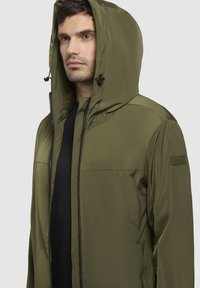 Olivenfarbene Outdoor-Jacke mit durchgehendem Reißverschluss, verstellbarem Kapuzen und Seitentaschen. Glattes, wasserabweisendes Material mit matter Oberfläche.
