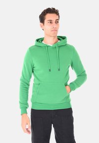 Giovane uomo che indossa un felpa verde con tasca frontale e pantaloni neri, in piedi con una mano in tasca, che guarda di lato.