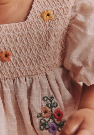 Enfant portant une robe rose clair avec des manches bouffantes, avec un corsage froncé et des fleurs brodées colorées sur la poitrine et la jupe.