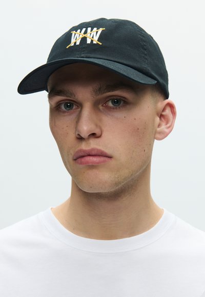 Μαύρο καπέλο με κεντημένο λευκό λογότυπο και κίτρινα διακοσμητικά, με καμπυλωτή γείσο και ρυθμιζόμενο λουράκι στο πίσω μέρος. Φορώντας με λευκό πουκάμισο.