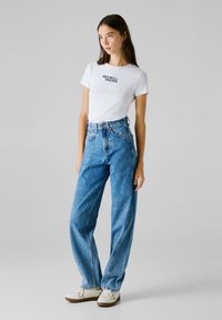 Joven mujer de pie, vistiendo una camiseta blanca de "Red Bull Racing", jeans de tiro alto de color azul y zapatillas blancas, frente a un fondo liso.