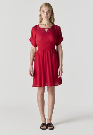 Mujer de pie mirando hacia adelante, vestida con un vestido rojo hasta la rodilla con mangas cortas y sandalias negras sin tacón, sobre un fondo claro y sencillo.