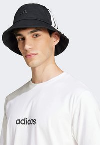 Zwarte bucket hat met witte gestreepte accenten en een geborduurd logo op de voorkant; gedragen met een eenvoudig wit T-shirt waarop een merknaam staat.