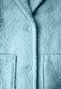 Cappotto azzurro chiaro con texture, realizzato in un morbido tessuto riccio, con ampie tasche frontali e chiusura con bottoni in plastica trasparente.