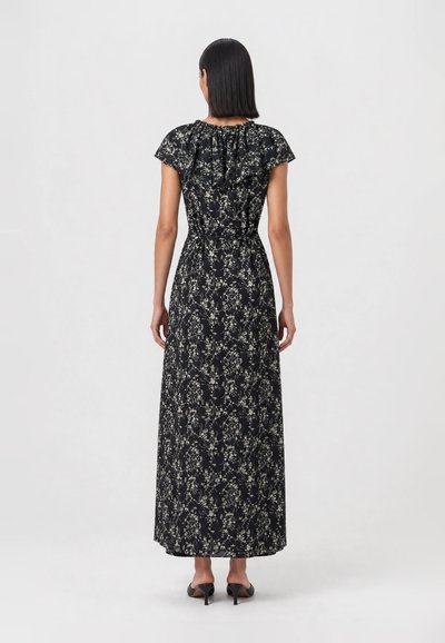 Woman standing facing away wearing a black floral pattern maxi dress with short sleeves and black heeled shoes.