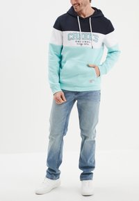 Kaptur z akwamaryny, granatu i bieli, z kieszenią z przodu, sznurkami i napisem logo. W zestawieniu z jasnoniebieskimi dżinsami i białymi sneakersami.
