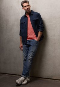 Homme appuyé contre un mur en béton, portant une veste bleu marine, une chemise corail, un jean bleu et des baskets blanches, les mains dans les poches.