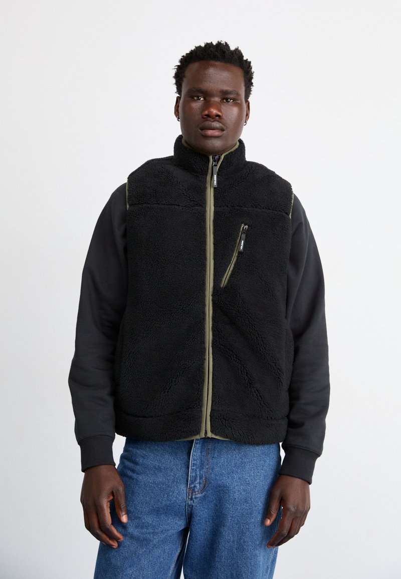 Gilet en polaire noir avec col haut, accents de fermeture éclair verts et poche latérale. Porté sur une chemise noire à manches longues et un jean bleu.