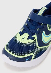 Basket de sport en mailles bleu et vert néon avec un dessus texturé, lacets et une sangle en Velcro ; présente une semelle blanche et des accents de logo en relief.