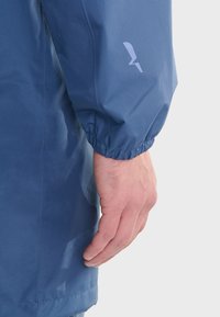 Blue waterproof jacket with elastic cuff, smooth texture, and a subtle logo detail on the sleeve. Close-up of sleeve and hand.