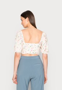 Top cropped floral com mangas bufantes em cores pastéis; apresenta uma parte de trás baixa e um tecido texturizado. Combinado com calças azuis de cintura alta.