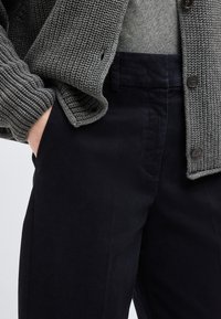 Άτομο που φοράει σκούρο ναυτικό μπλε παντελόνι και γκρι πλεκτή ζακέτα με κουμπιά, το χέρι του είναι βαλμένο στην τσέπη.