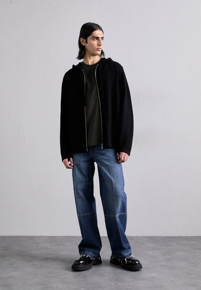 Schwarze Zip-Jacke mit Kapuze, olivgrünes Shirt darunter, blaue weite Jeans mit zusammengesetztem Denim, schwarze Plateauschuhe, vor einem neutralen Hintergrund stehend.