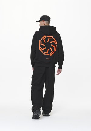 Svart hettegenser med et stort oransje grafisk design på ryggen, kombinert med svarte cargobukser og svarte sneakers. Enkel, funksjonell design.
