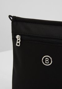 Svart nylonväska med en silverdragkedja som har en distinkt "B"-dragare och en cirkulär logotyp med samma bokstav. Slät yta, enkelt design.