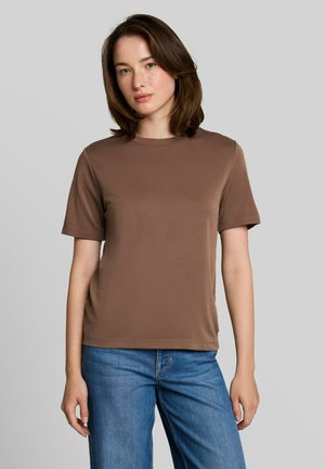 Junge Frau mit mittellangem braunen Haar, die ein schlichtes braunes Kurzarmshirt und blaue Jeans vor einem grauen Hintergrund trägt.