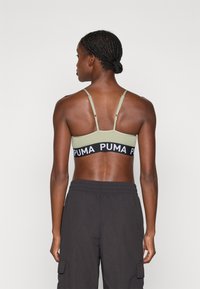 Puma sportski grudnjak u maslinasto zelenoj boji s crnim remenom na kojem je logo "PUMA". Podešive naramenice i glatka tekstura, dizajniran za udobnost i potporu.