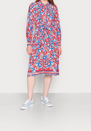 Blomstrende kjole med røde, blå og hvide farver, lange ærmer og en tætsiddende talje. Parret med lyseblå sneakers med hvide såler.