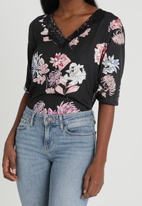 Blouse noire à motif floral avec un col en V bordé de dentelle et des manches à coudes, associée à un jean taille haute bleu clair.