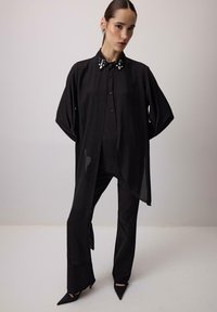 Czarna, oversize koszula z ozdobnym kołnierzem, z zapięciem na guziki z przodu. Stylizowana z dopasowanymi czarnymi spodniami i spiczastymi czarnymi szpilkami.