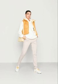 Colete acolchoado mostarda sobre um hoodie branco, calças bege claras e ténis brancos com detalhes amarelos. O modelo está de pé com uma mão na anca.