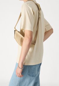 Bolso cruzado beige hecho de un tejido suave, con forma curva, correa ajustable y cierre de cremallera. Llevado sobre una blusa de color claro.
