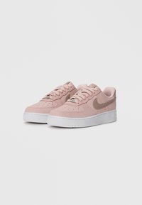 Um par de ténis Nike Air Force 1 cor de rosa claro com tecido texturizado e logótipos beige do swoosh, solas brancas e atacadores num fundo branco liso.