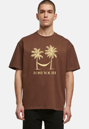 Mann in braunem, oversized T-Shirt mit gelben Palmen, Grafiken einer Hängematte und dem Text "VERLORENES JUGEND", der vor einem weißen Hintergrund steht.