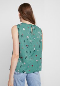 Blusa verde senza maniche con motivo floreale, caratterizzata da un dettaglio con bottone a chiave sulla schiena e una vestibilità rilassata, abbinata a jeans azzurri chiari.